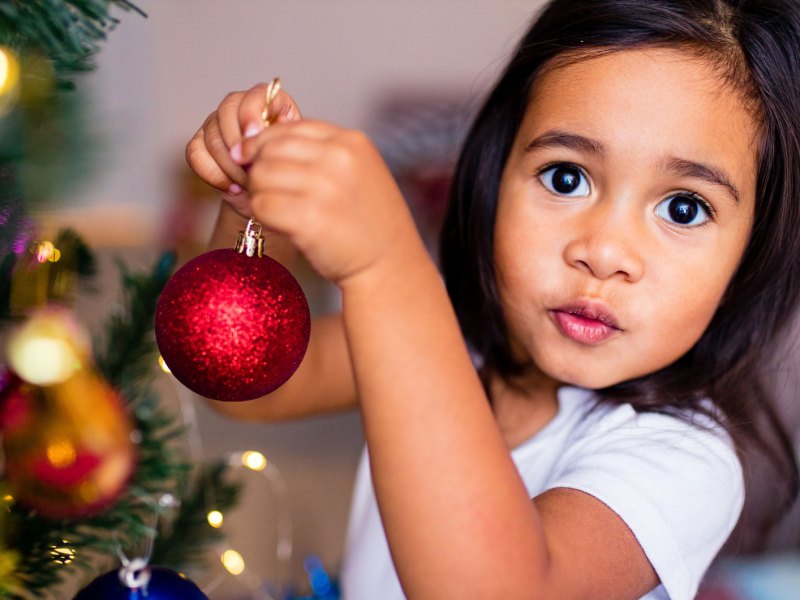 Nahaufnahme eines kleinen Mädchens, ca. 6 Jahre alt, das den Weihnachtsbaum mit Kugeln schmückt.