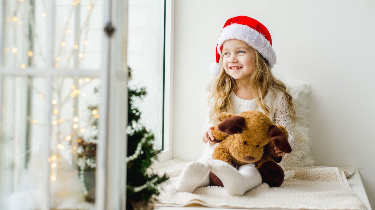 Mädchen, ca. 5 Jahre alt, sitzt mit einer Weihnachtsmannmütze am Fenster und schaut neugierig raus.