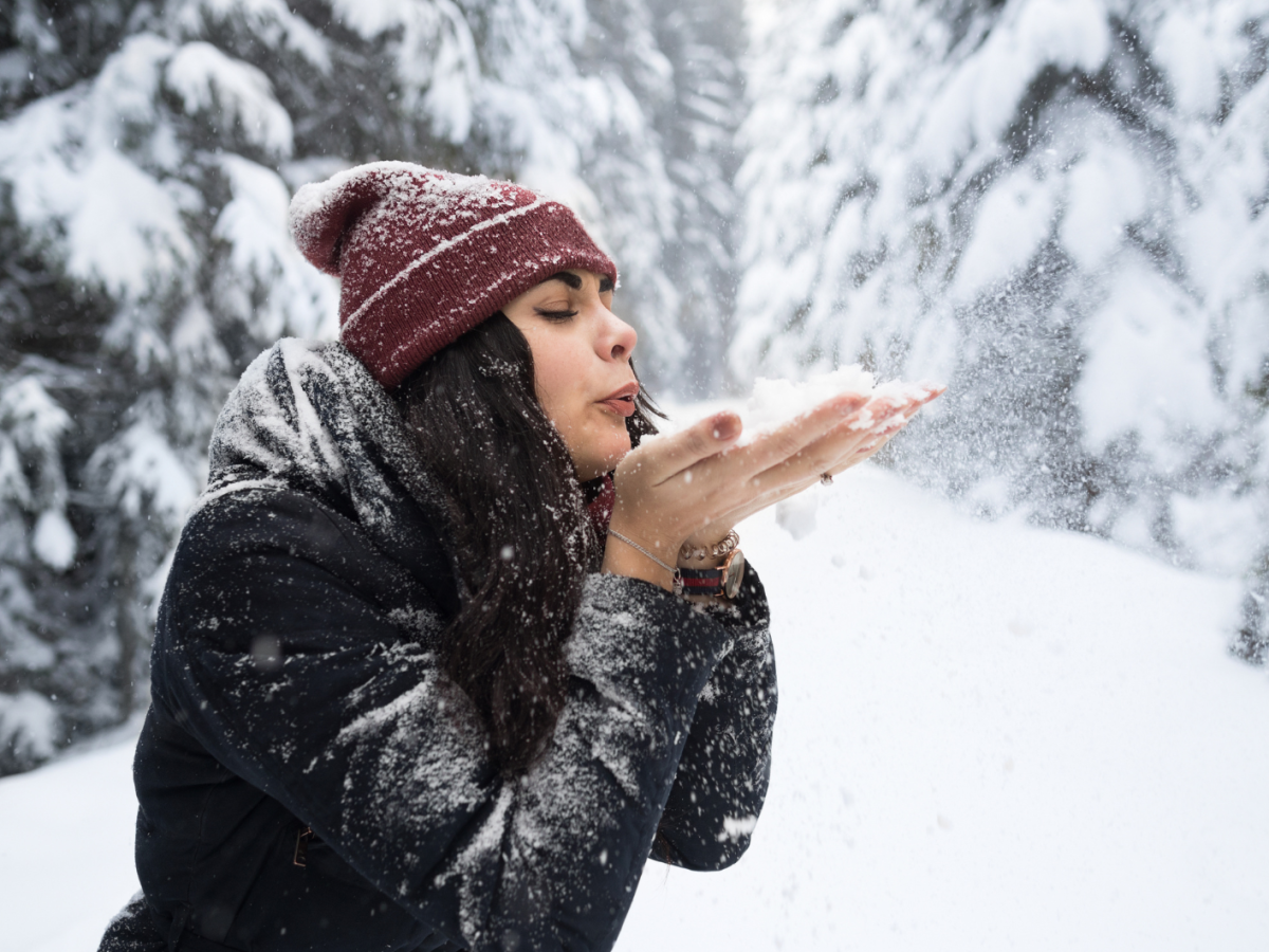 Junge Frau mit schwarzem Haar und roter Mütze pustet Schnee aus ihren Händen in einem verschneiten Wald voller Tannen.