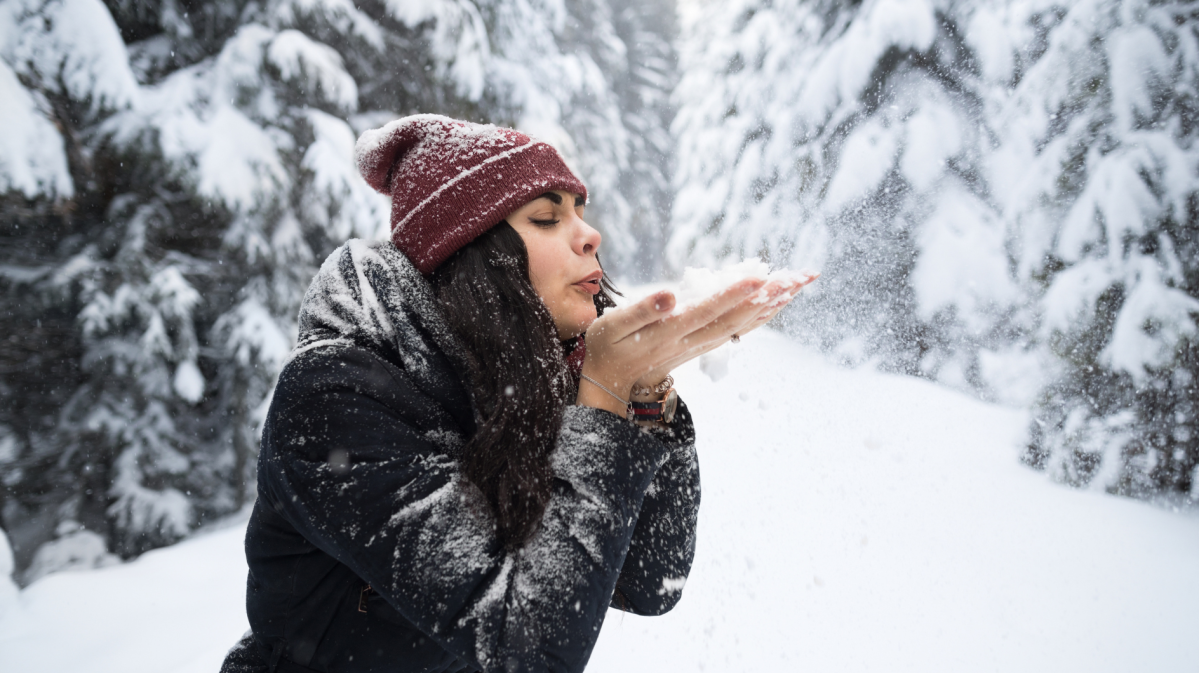 Junge Frau mit schwarzem Haar und roter Mütze pustet Schnee aus ihren Händen in einem verschneiten Wald voller Tannen.