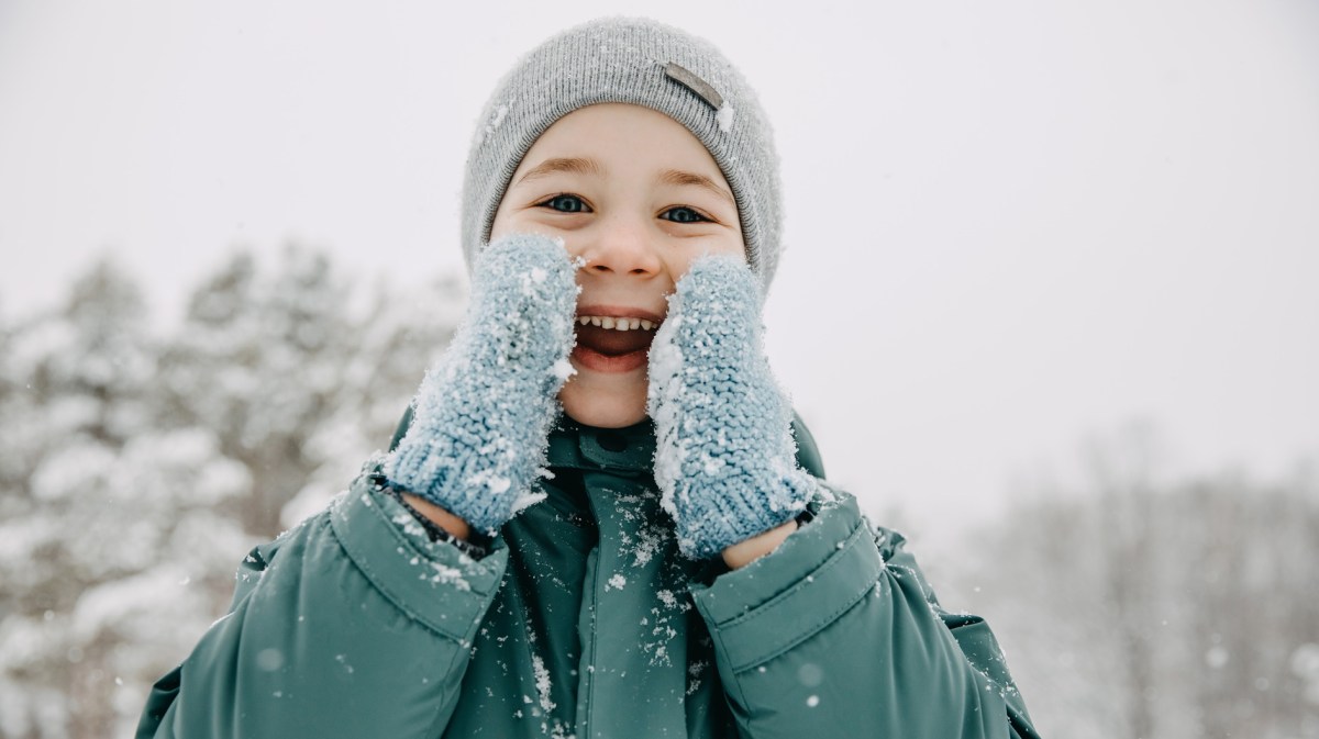 Kind an einem verschneiten Wintertag strahlt in die Kamera und hält sich seine Hände in Handschuhen ans Gesicht.