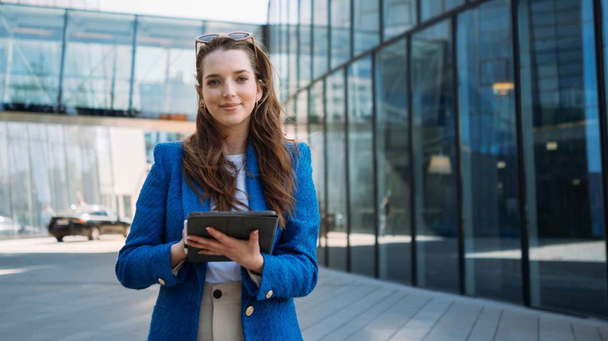 Frau mit braunen Haaren und einem blauen Blazer steht vor einem Bürogebäude und hält ein Tablet in den Händen.