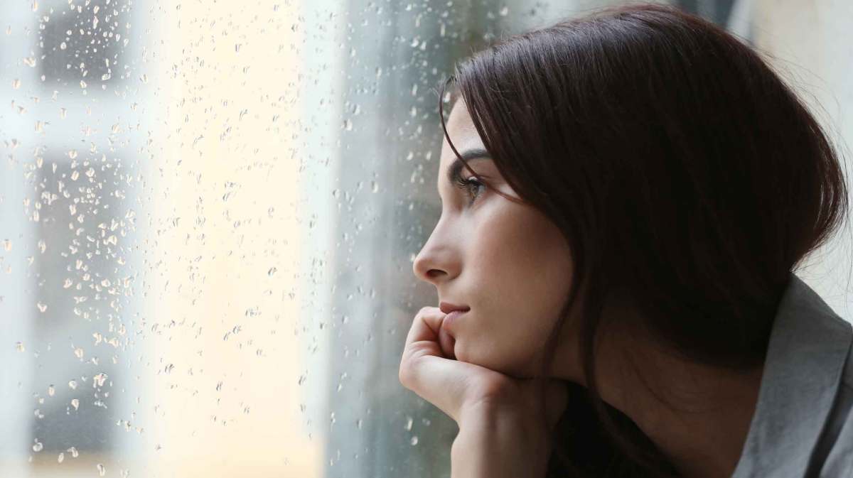 Eine dunkelhaarige Frau guckt im Januar traurig aus dem Fenster, während es draußen regnet.