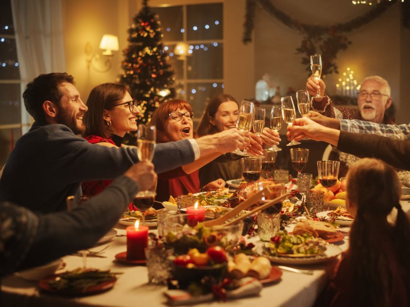 Familie und Freunde sitzen an einem festlich gedeckten Tisch, im Hintergrund leuchtet der Weihnachtsbaum und alle prosten sich zu.