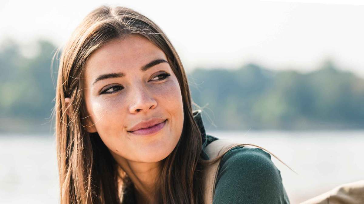 Junge Frau mit glatte dunklen Haaren sitzt draußen am Wasser und blickt freundlich zur Seite.