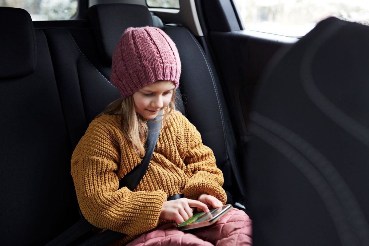 Mädchen mit rosa Mütze und gelbem Pullover sitzt angeschnallt im Auto und schaut aufs Handy