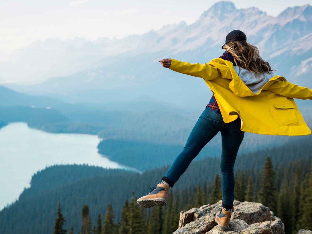 Frau steht waghalsig auf einem Felsen mit einem Bein vor einer Berglandschaft.