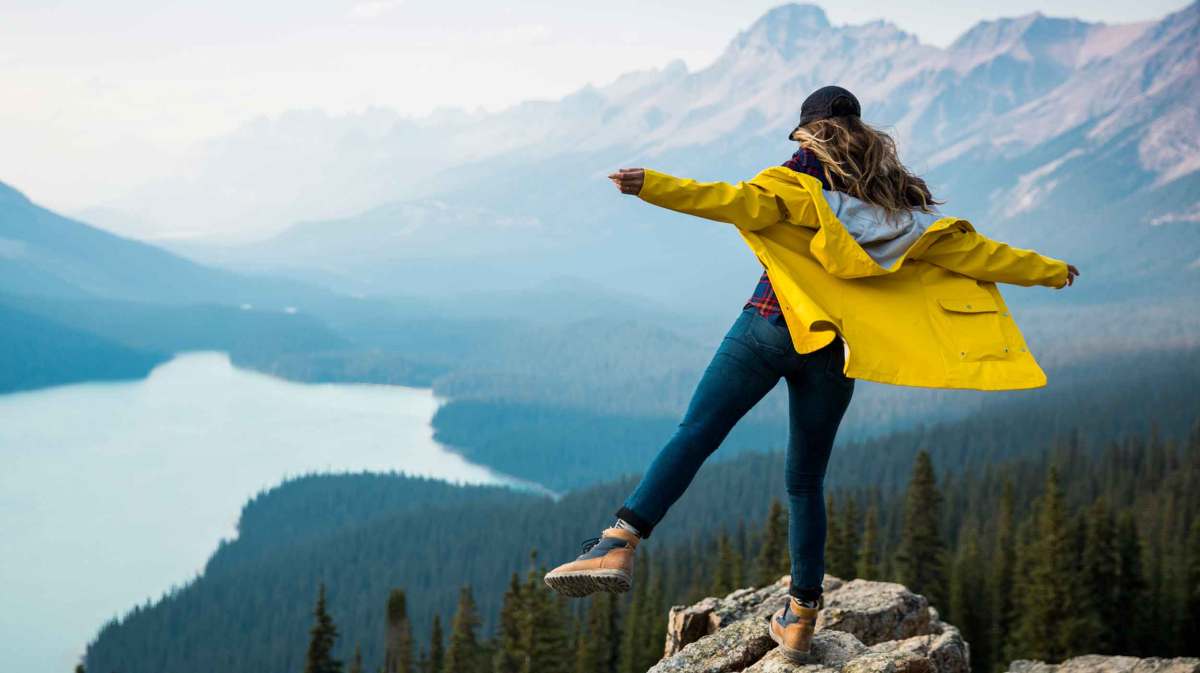 Frau steht waghalsig auf einem Felsen mit einem Bein vor einer Berglandschaft.