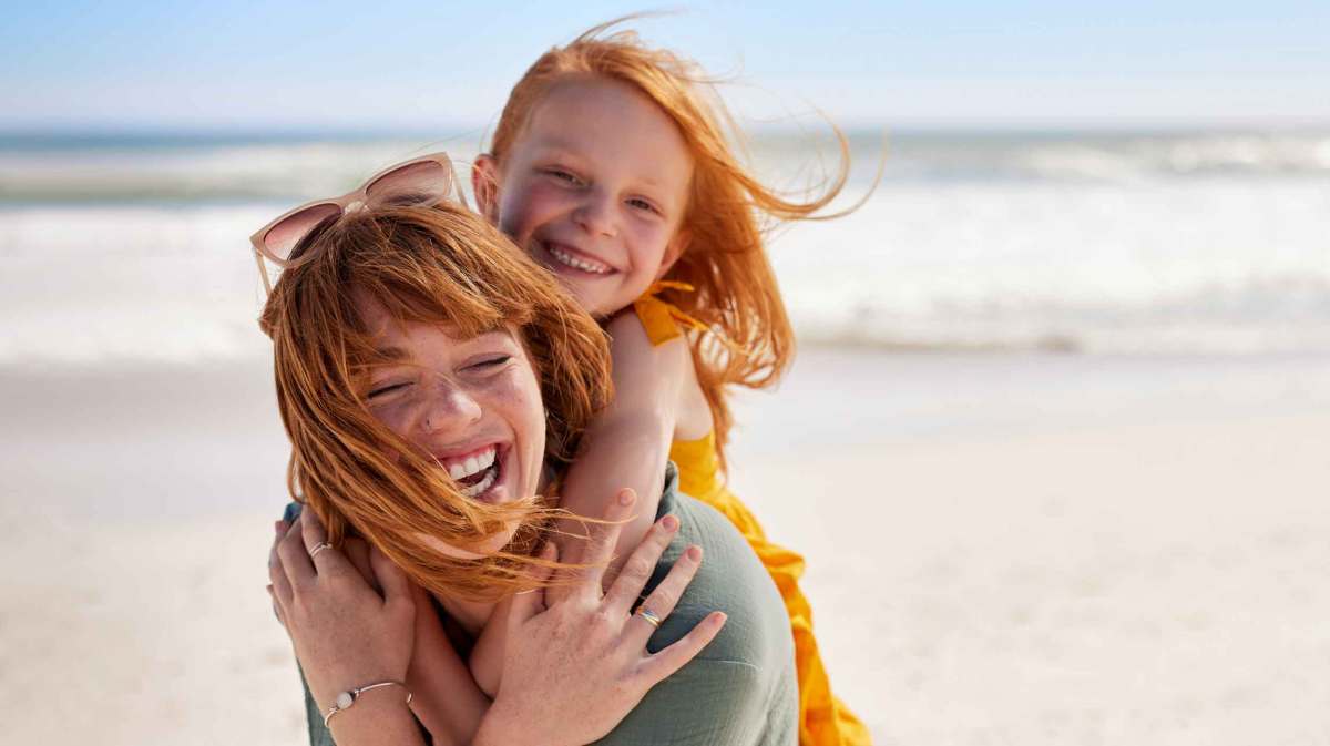 Eine lachende rothaarige Frau trägt ein rothaariges Mädchen in gelbem Kleid am sonnigen Strand, beide wirken ausgelassen.