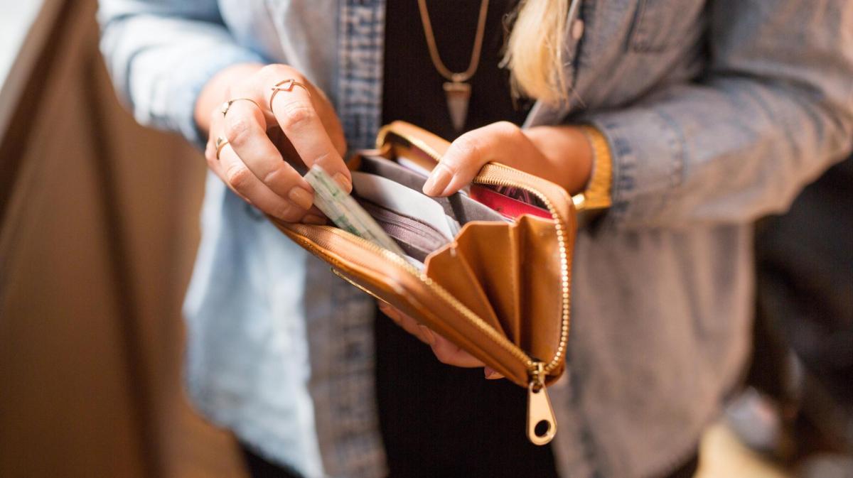 Eine Frau in Jeanshemd und schwarzem Oberteil nimmt einen Tampon aus ihrer braunen Geldbörse, ihre Hände tragen goldene Ringe.