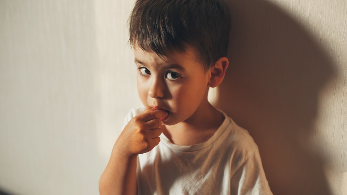 Junge, ca. 4 Jahre, steht vor einer weißen Wand und schaut misstrauisch in die Kamera.