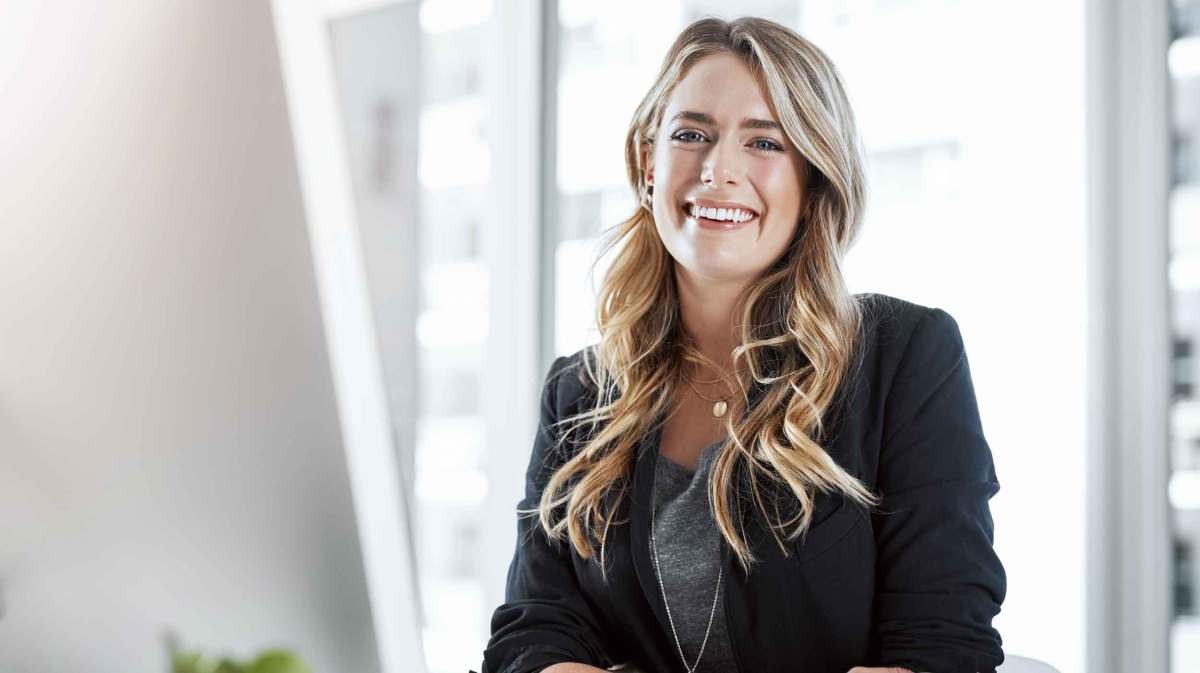 Eine junge Frau mit langen blond gewellten Haaren und schwarzem Blazer lächelt freundlich in einem modernen, lichtdurchfluteten Büro.