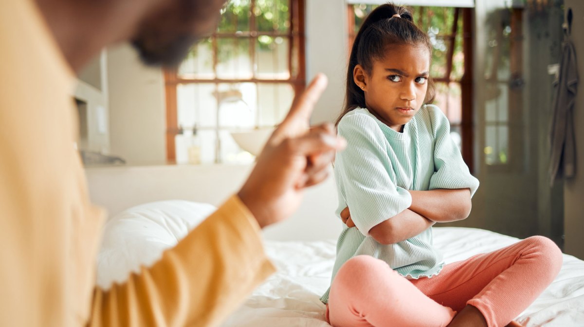 Wütendes Mädchen sitzt auf ihrem Bett, verschränkt die Arme und schaut ihren Vater böse an.