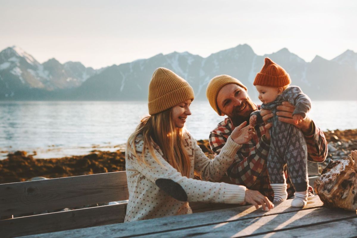 Eltern und Kleinkind mit warmer Kleidung sitzen draußen am Tisch vor einem Bergsee