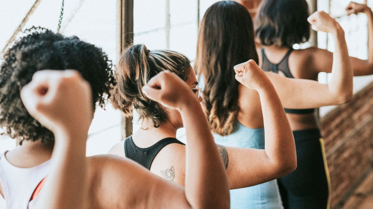 Frauen in Fitness-Bekleidung stehen mit dem Rücken zur Kamera und heben Arm mit geballter Faust.