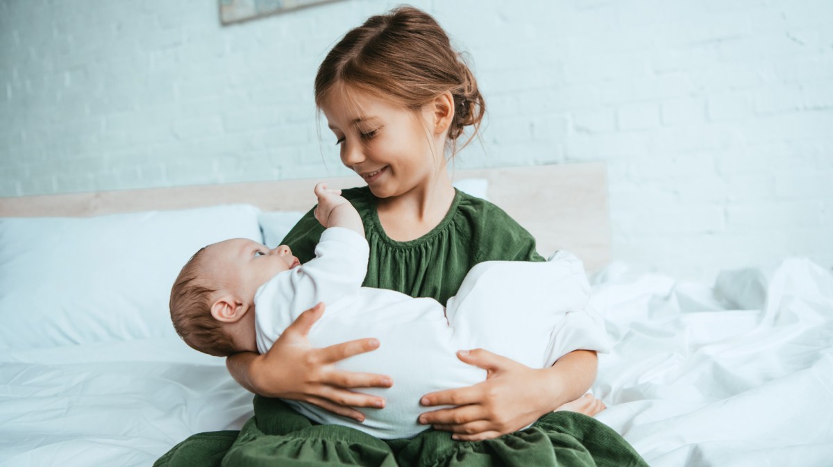 Glückliches Mädchen hält ihre kleine Baby-Schwester im Arm, während sie auf einem weißen Bett sitzt.