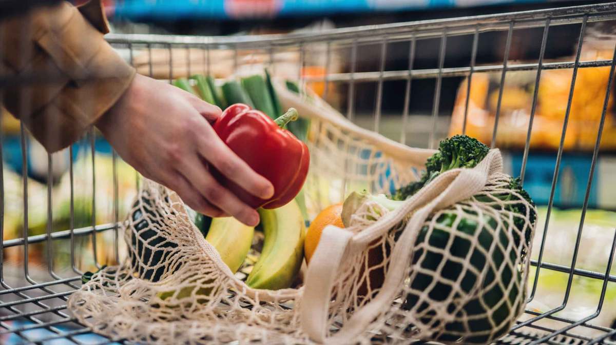 Eine Hand legt eine rote Paprika in einen Einkaufswagen, in dem in einem Netzbeutel Obst und Gemüse liegen, darunter Bananen und Brokkoli.