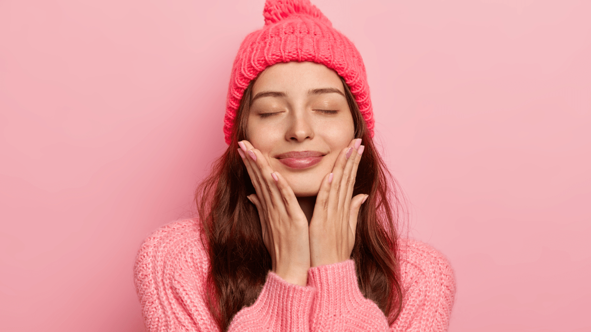 Junge Frau mit geschlossenen Augen lächelt entspannt, trägt eine pinke Strickmütze und Pullover vor rosa Hintergrund.