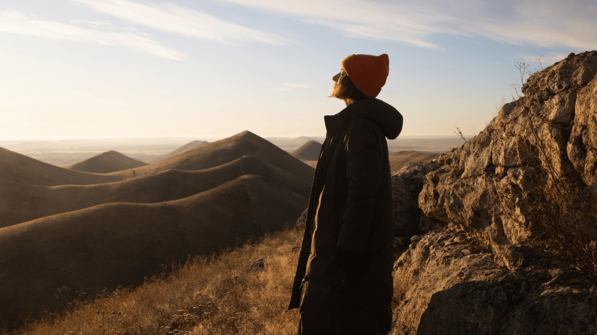 Eine Person mit orangefarbener Mütze und langem Mantel steht bei Sonnenuntergang auf einem Felsvorsprung und blickt über hügelige Landschaft.