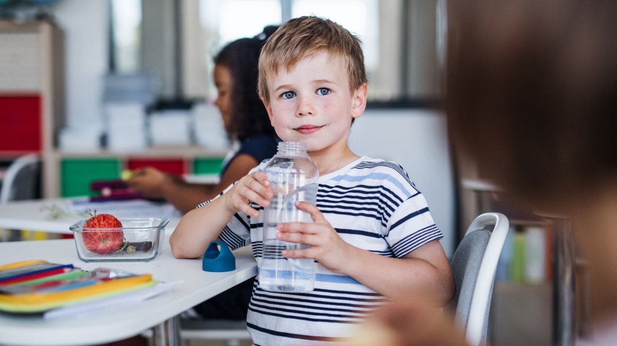 Schuljunge im Klassenraum hält eine große Wasserflasche in seinen Händen und hat sich seitlich zur Kamera gedreht.