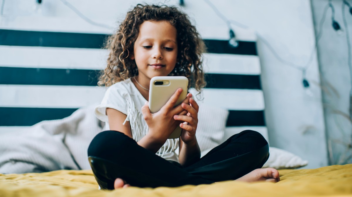 Mädchen, ca. 8 Jahre alt, sitzt mit ihrem Handy im Schneidersitz auf dem Bett. Es lächelt den Bildschirm an.