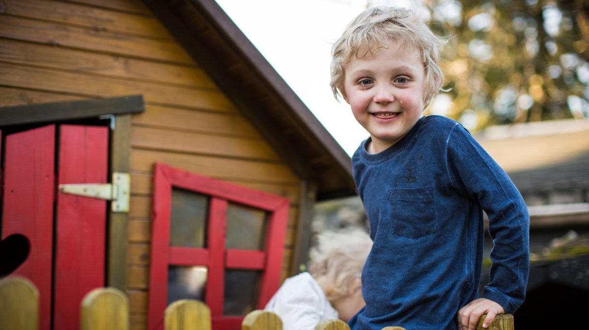 Junge, ca 5 Jahre alt, steht vor einem Spielhaus aus Holz. Im Hintergrund ist ein weiteres, jüngeres Kind zu sehen.