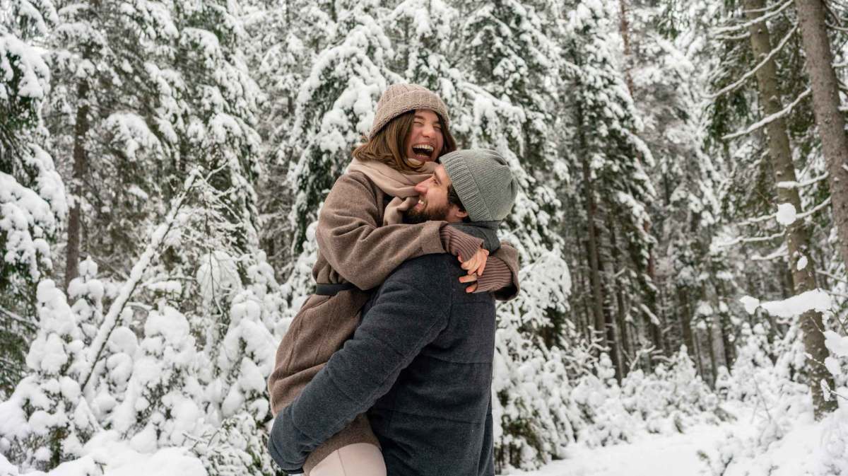 Mann hebt fröhliche Frau im braunen Mantel hoch, beide lachen herzlich in einem verschneiten Nadelwald.