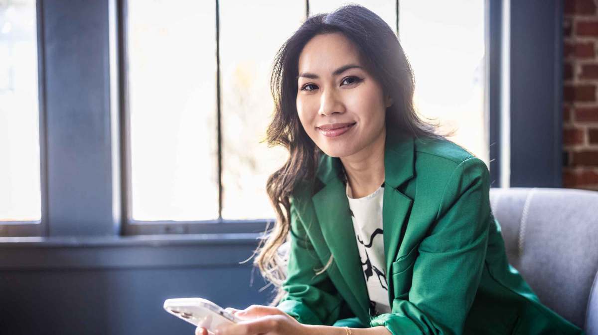 Junge dunkelhaarige Frau mit einem grünen Blazer und einem modernen Look sitzt entspannt bei der Arbeit auf einer Sitzgelegenheit und lächelt, während sie ihr Handy in der Hand hält.