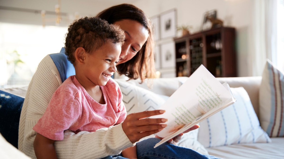 Mutter und Sohn, ca. 4 Jahre alt, sitzen auf dem Sofa und lesen gemeinsam ein Buch. Das Kind lacht.