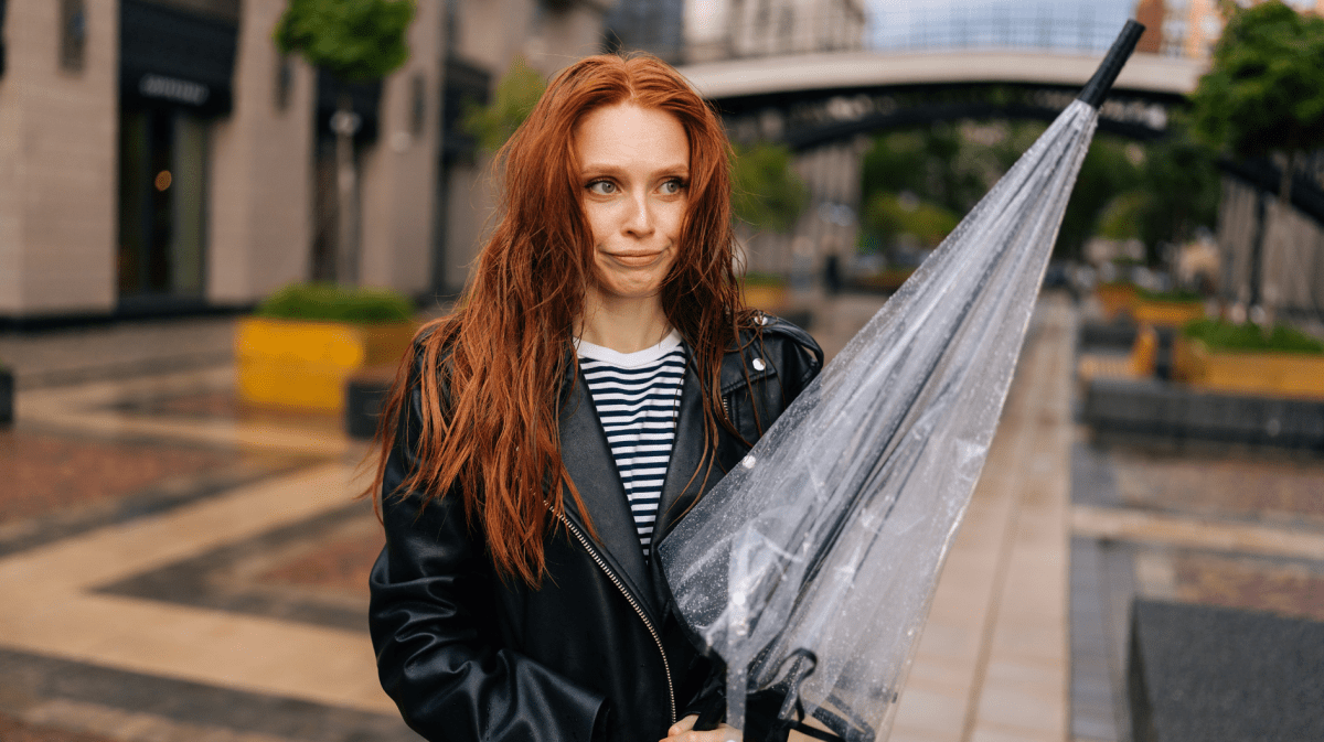 Frau mit nassen und durchwuschelten Haaren, die einen Regenschirm in der Hand hält und schmollend zur Seite schaut.