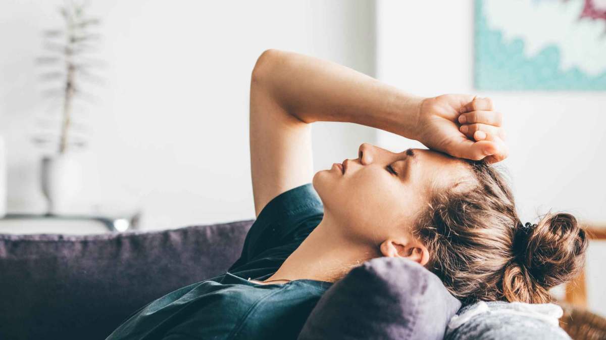 Eine junge Frau mit dunklem Dutt lehnt erschöpft auf einem Sofa, ihre Augen geschlossen und die Hand an die Stirn gelegt.