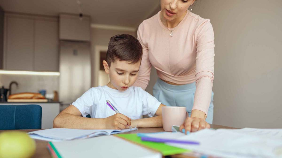 Ein Grundschulkind arbeitet mit Stift an den Hausaufgaben, neben ihm steht eine Frau und zeigt auf ein Blatt.