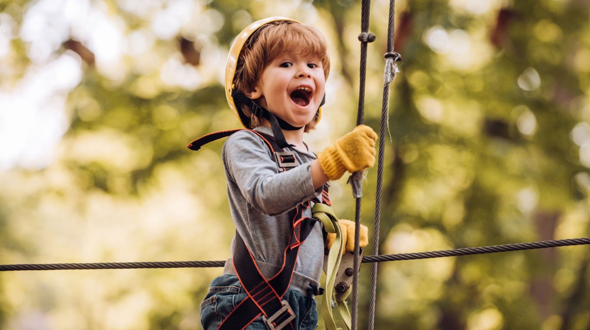 Junge, ca. 6 Jahre alt, in voller Montur auf einem Seil in einem Kletterwald.