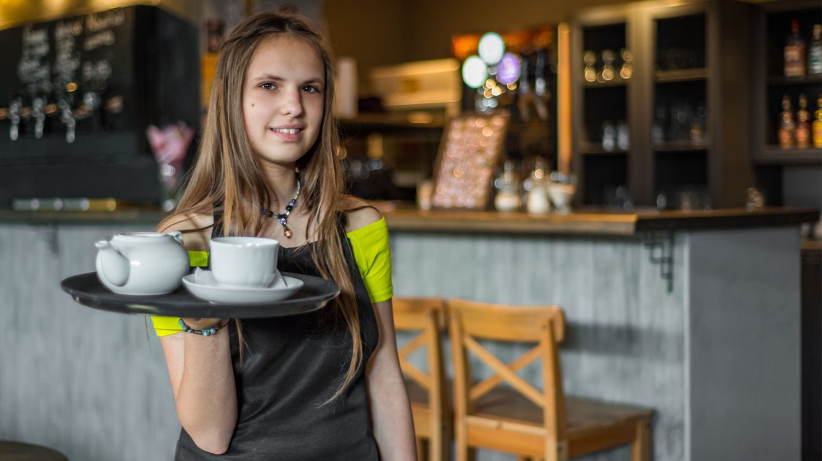 Mädchen im Teenageralter in einem Kaffee. Es trägt eine Schürze und ein Tablett.