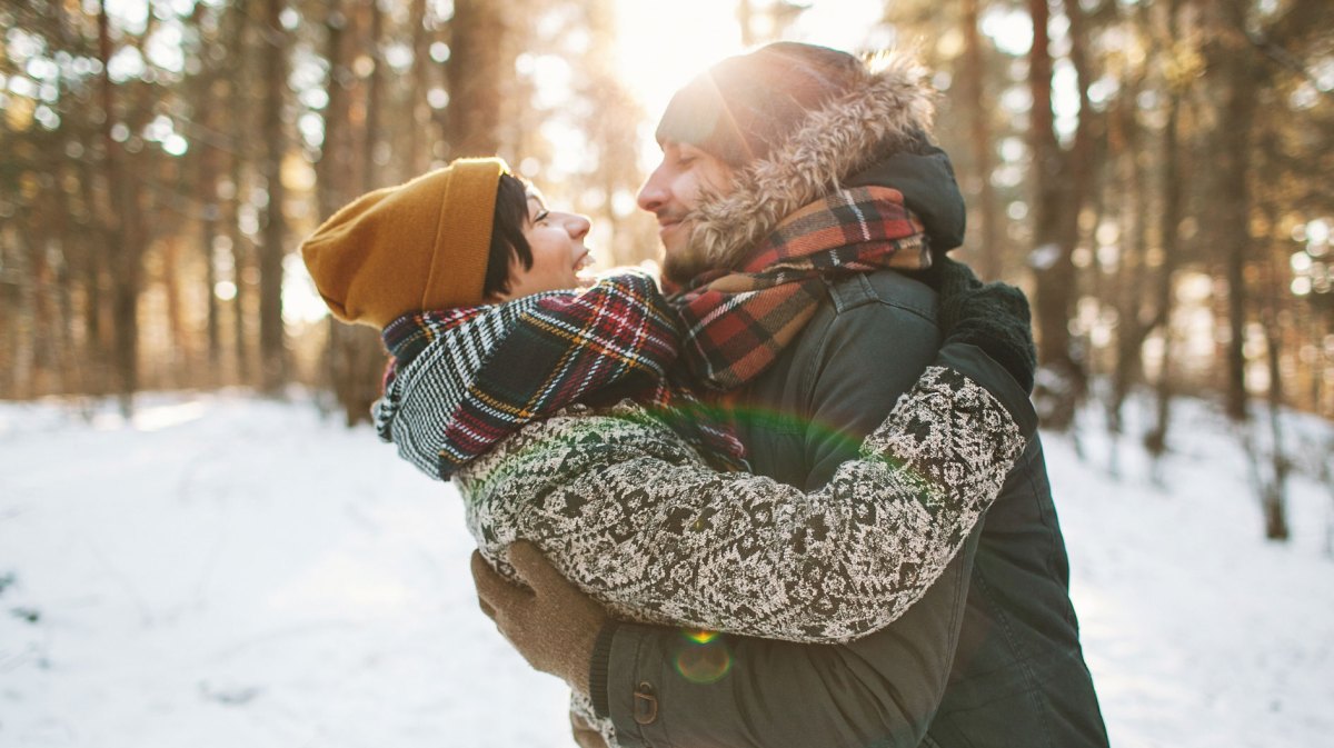 Mann und Frau in Winterbekleidung lachen und umarmen sich in verschneitem Wald