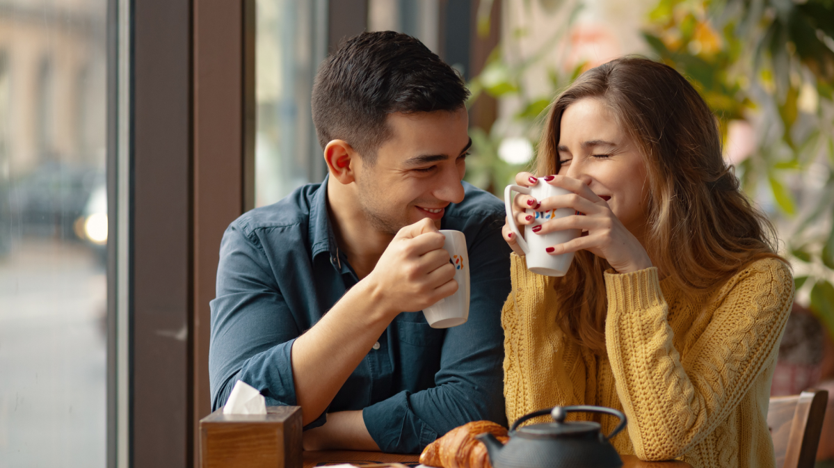 Frau und Mann mit Tassen in der Hand sitzen lachend an einem Tisch in einem Café