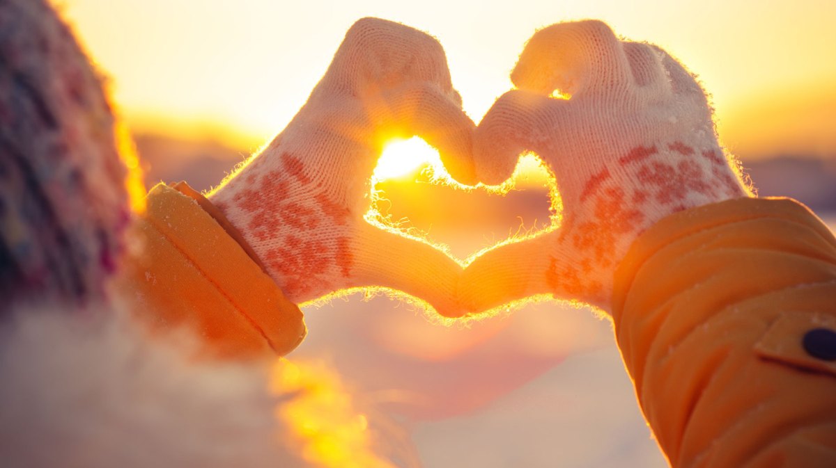 Person in Winterbekleidung formt ein Herz mit ihren Händen in winterlicher Landschaft bei Sonnenuntergang