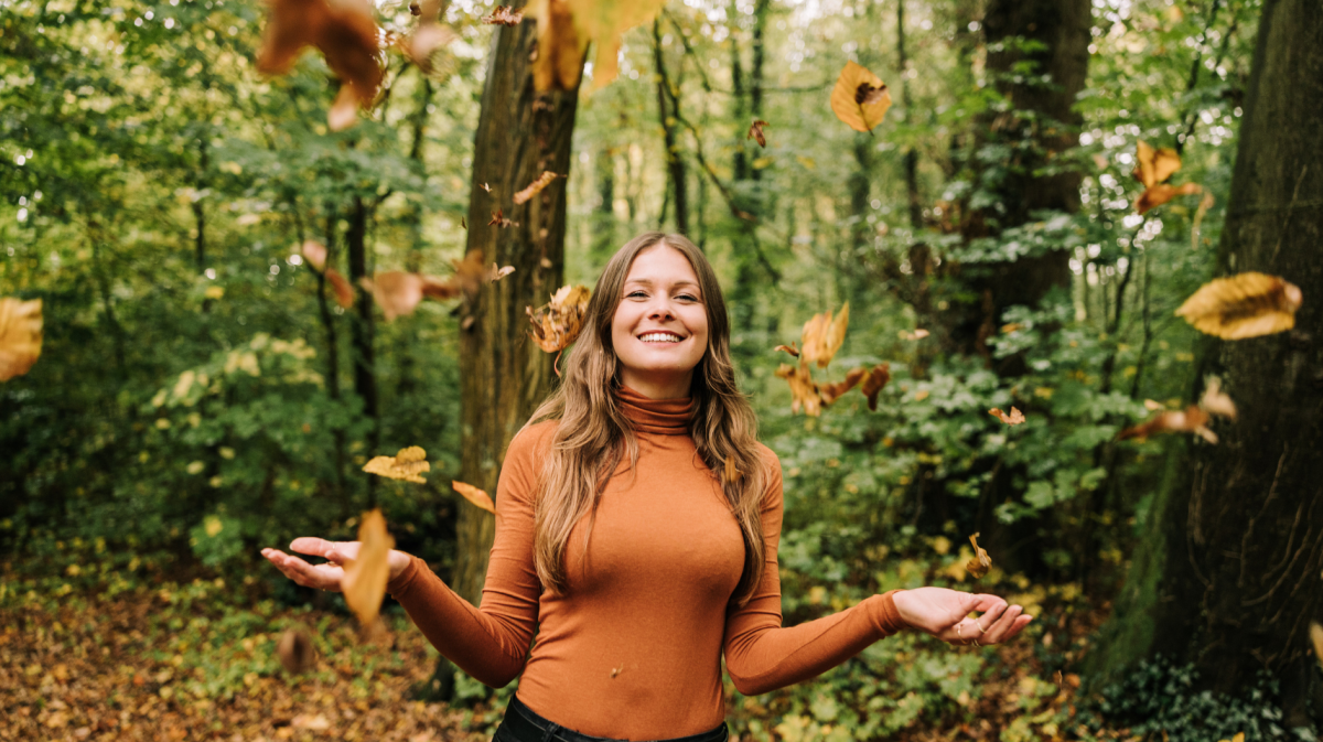 Frau im Wald, die lacht und Blätter in die Luft wirft.