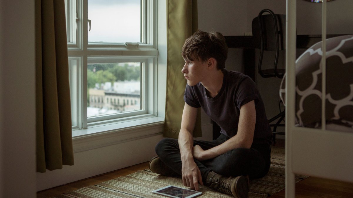 Junge, ca. 15 Jahre, sitzt im Schneidersitz auf dem Boden, vor ihm ein Tablet auf dem Boden und schaut melancholisch aus dem Fenster.