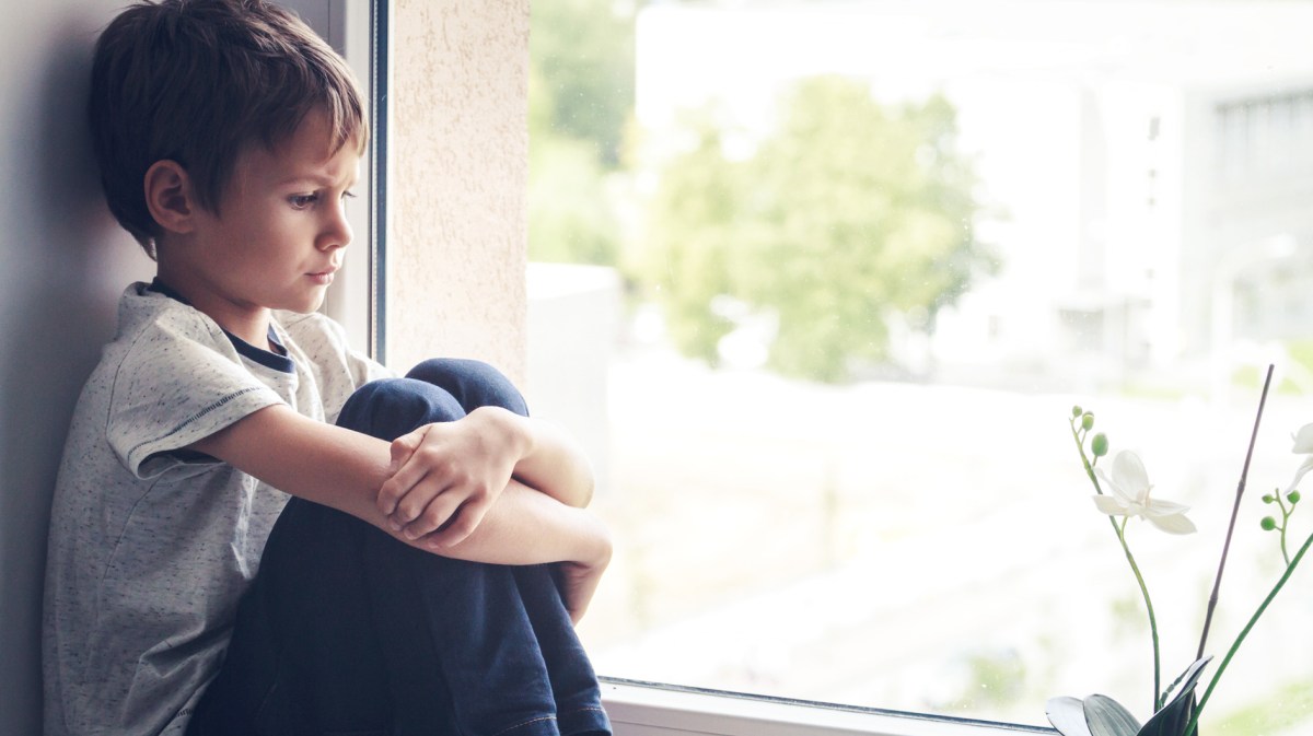 Trauriger Junge, ca. 8 Jahre alt, sitzt auf dem Fensterbrett und hält seine Beine mit seinen Armen umschlungen.