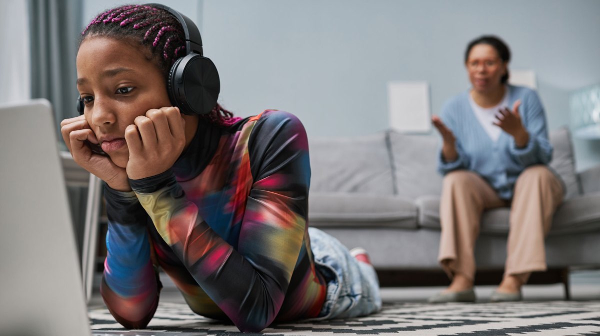 Teenager-Mädchen liegt auf dem Fußboden, trägt Kopfhörer und schaut etwas auf ihrem Laptop. Die Mutter sitzt im Hintergrund auf der Couch und versucht, ihrer Tochter etwas zu erklären.