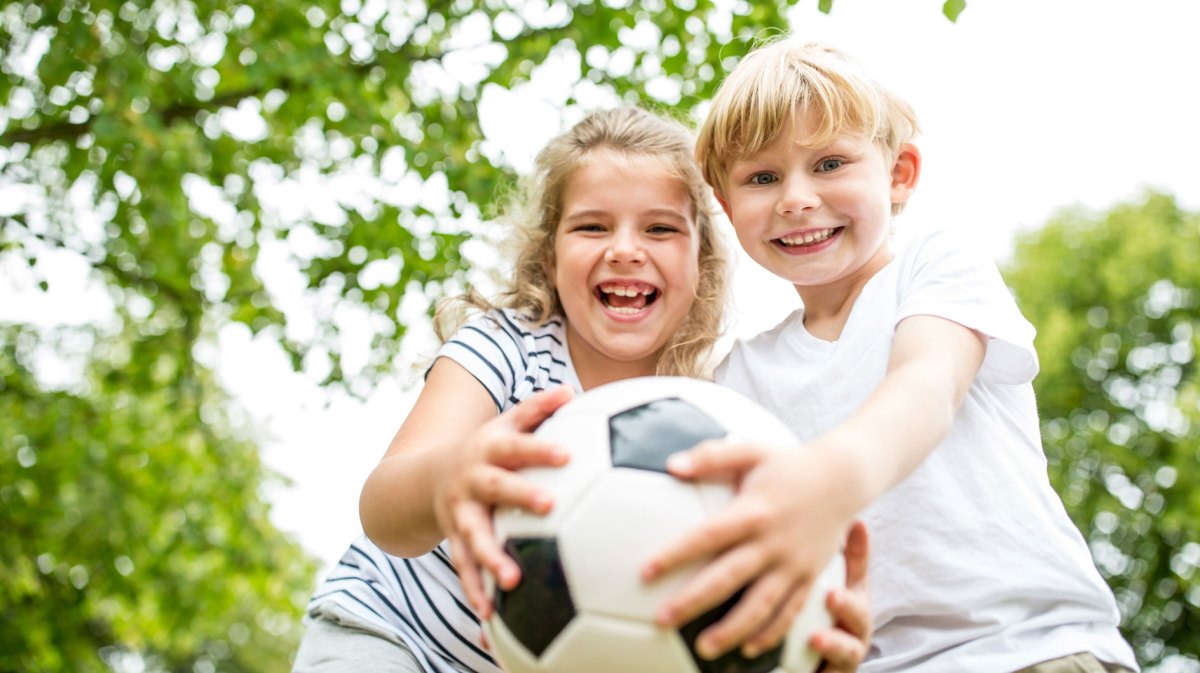 Geschwister halten gemeinsam einen Fußball fest und strahlen dabei in die Kamera.