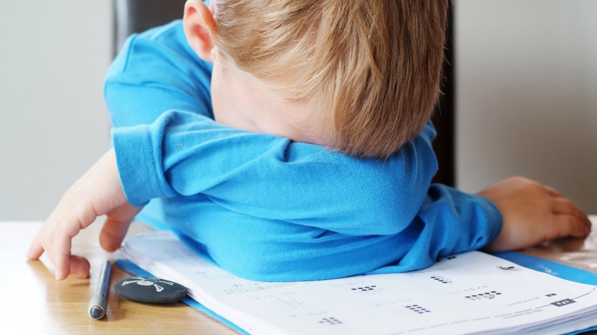 Junge im Grundschulalter hat seinen Kopf über ein paar Mathematikaufgaben auf seinen verschränkten Armen auf dem Schreibtisch abgelegt.