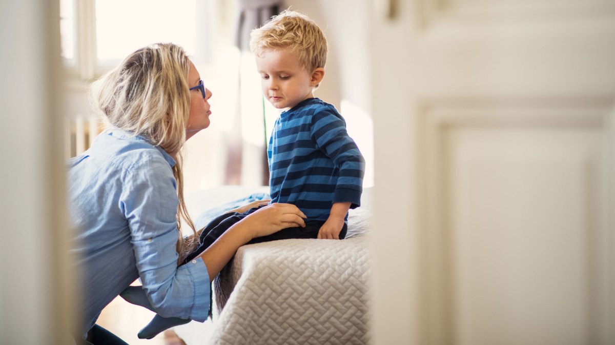 Mutter redet ernst mit ihrem Sohn, ca. 4 Jahre alt. Er sitzt auf dem Bett, sie hockt davor und redet auf Augenhöhe mit ihm.