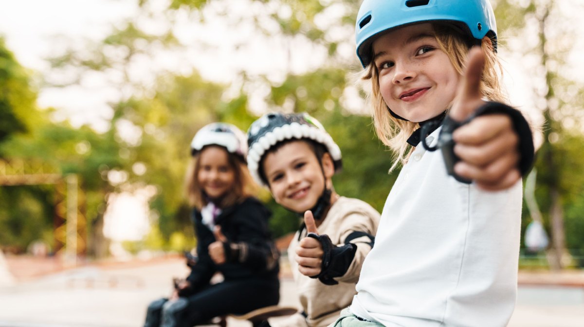 Drei Kinder sitzen in voller Sicherheitsmontur auf dem Skaterplatz und strecken den Daumen hoch Richtung Kamera.