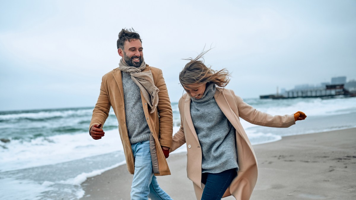 Glückliches Paar läuft lachend am Strand entlang im Winter.