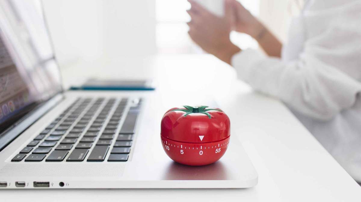 Eine Küchenuhr in Form einer Tomate steht auf einem Notebook. Im Hintergrund sitzt eine Frau mit einem Kaffee.