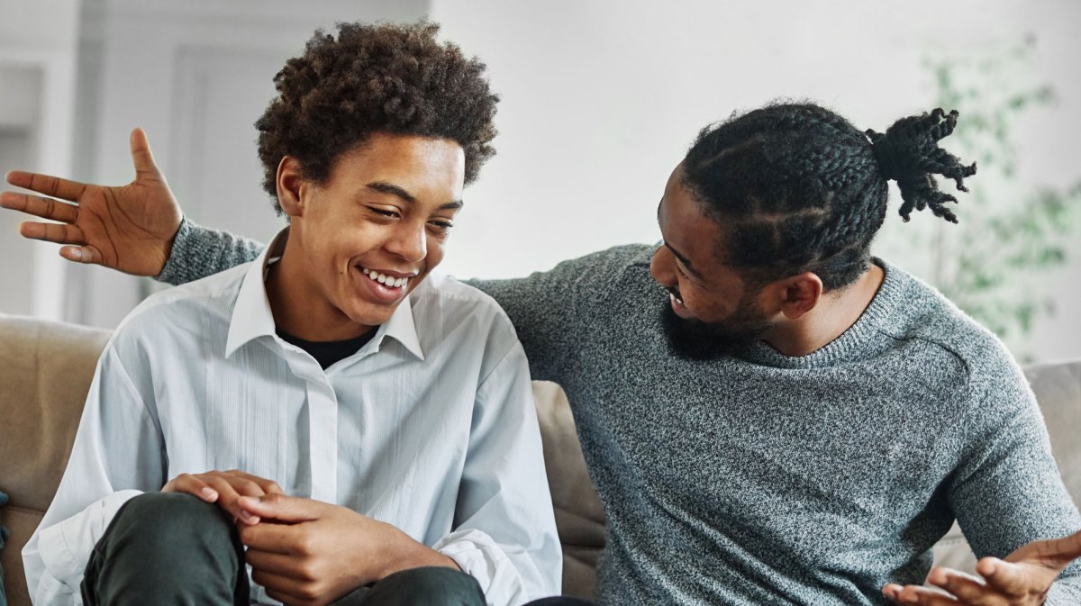 Vater und Sohn sitzen auf der Couch. Der Sohn lächelt erleichtert, leicht beschämt, der Vater freu sich in einer ausladenden Geste.