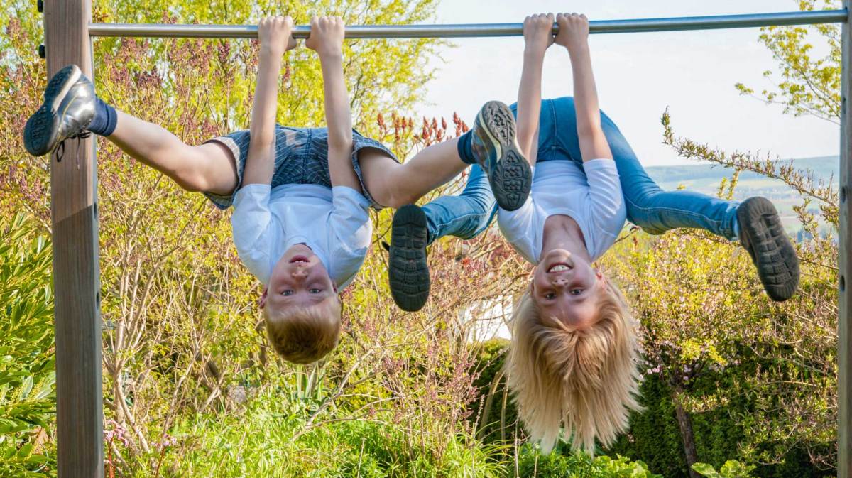 Zwei Kinder im Grundschulalter baumeln an einer Kletterstange mit dem Kopf nach unten.