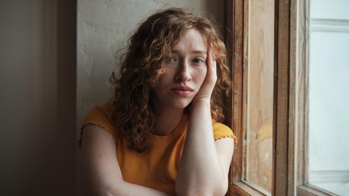 Junge Frau mit Locken sitzt am Fenster und guckt sehr traurig.